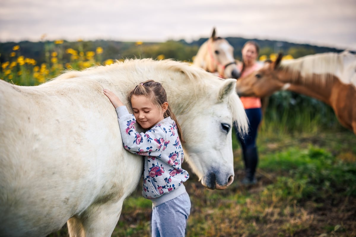 A young girl embracing a white pony on the farm while her mother watches